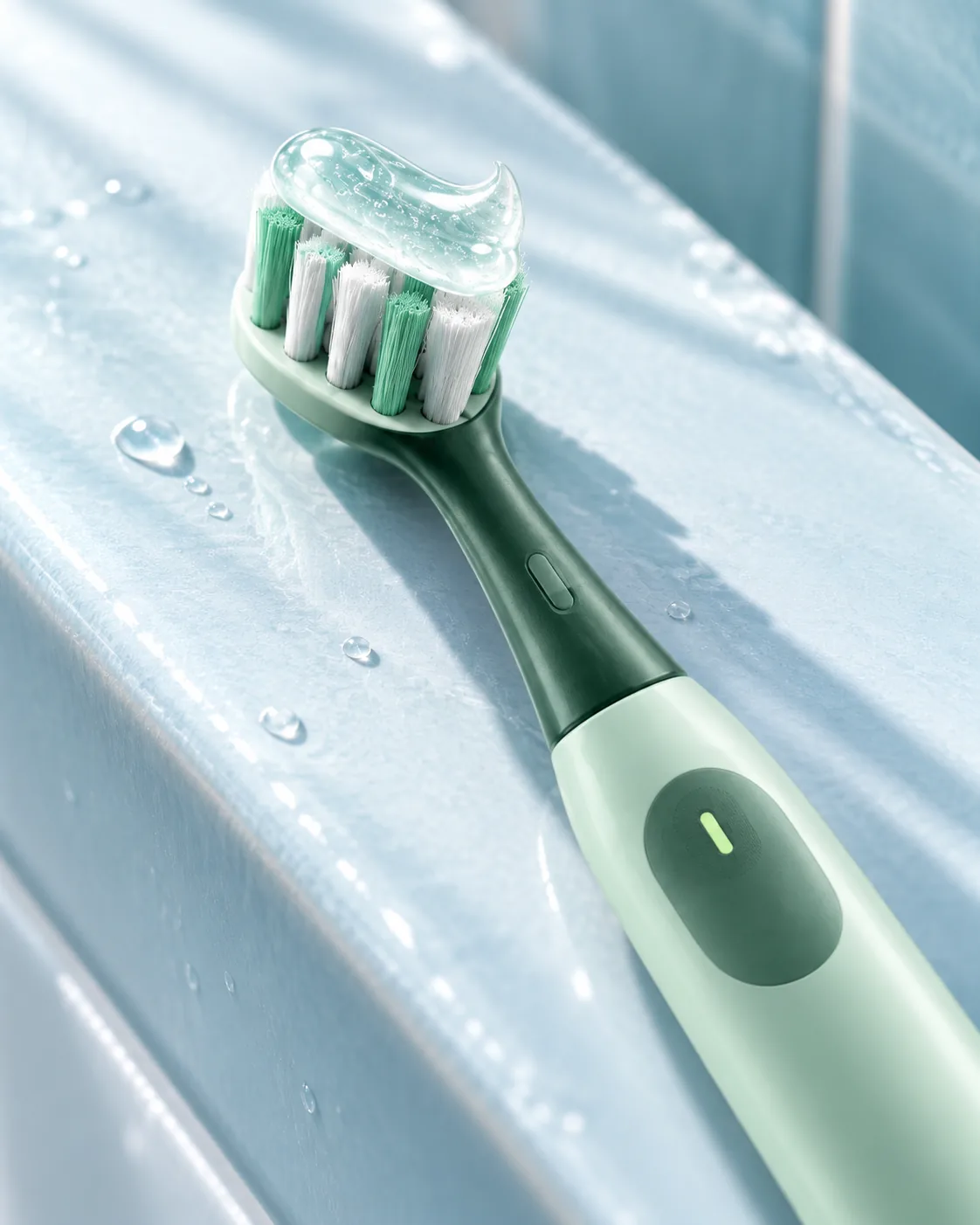 Close-up of a mint-green electric toothbrush with clear gel on its bristles on a pale blue ceramic ledge