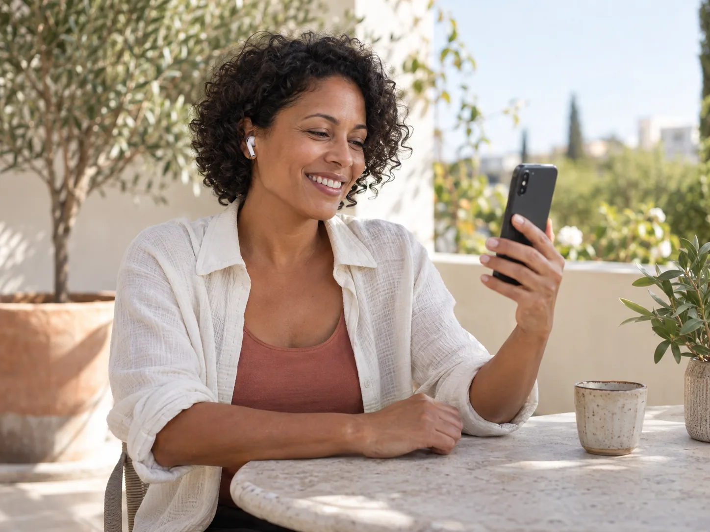 A woman with short curly hair and an earbud smiling at her phone on a sunlit terrace